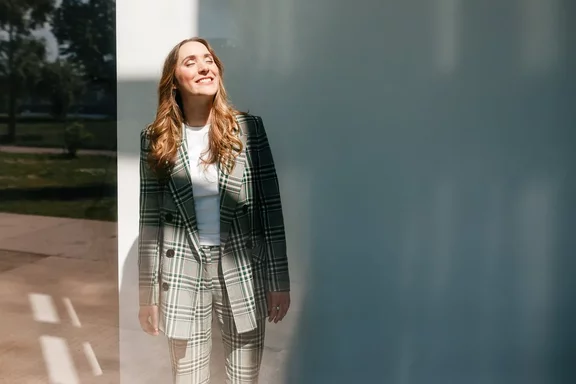Eine Frau mit langen Haaren, die einen quadratischen Blazer und eine Hose trägt, steht an einer weißen Wand und blickt mit einem freudigen Ausdruck nach oben.