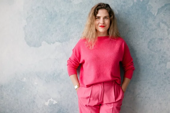 Eine Frau in einem leuchtend rosafarbenen Outfit steht vor einem farbigen blau-grauen Hintergrund und trägt eine Uhr und einen roten Lippenstift.