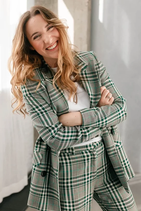 Eine Frau mit welligem braunem Haar trägt einen grün-weißen karierten Blazer und eine Hose, lächelt und posiert in einem Raum mit einem Fenster.