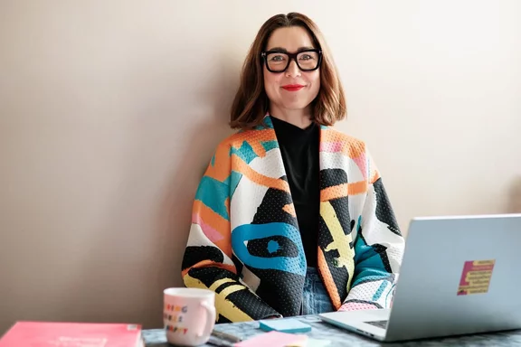 Eine Frau mit Brille und bunter Jacke sitzt an einem Tisch mit einem Laptop und einem Becher an einer beigen Wand.
